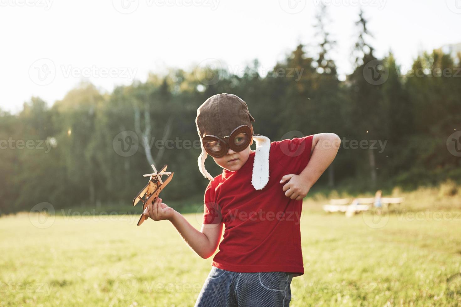 glückliches Kind im Pilotenhelm, das mit einem hölzernen Spielzeugflugzeug spielt und davon träumt, fliegen zu werden foto