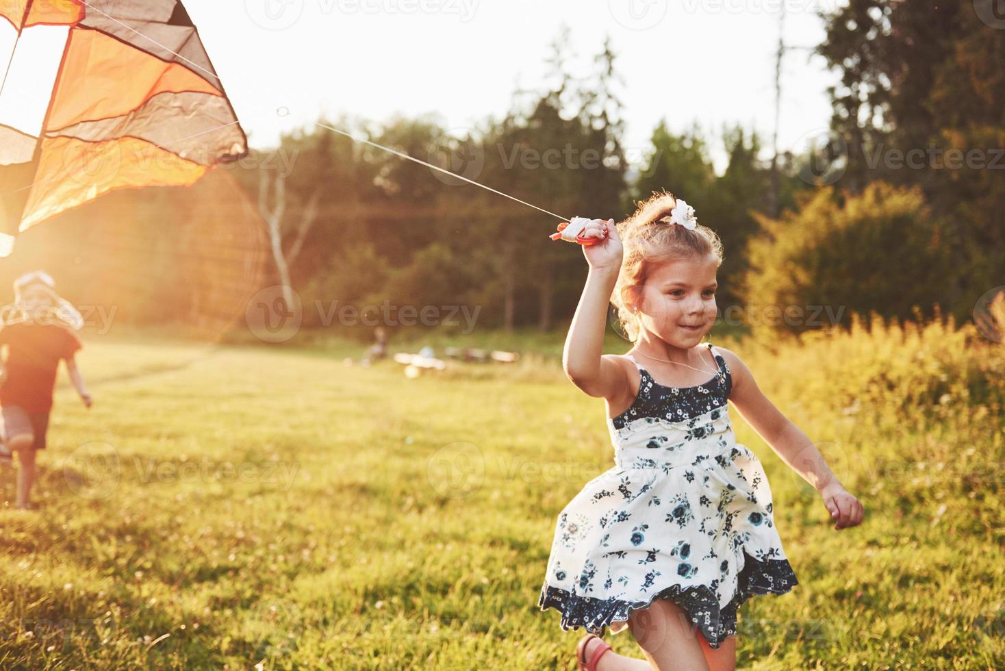 süßes kleines Mädchen mit langen Haaren, das an einem sonnigen Sommertag mit Drachen auf dem Feld läuft foto