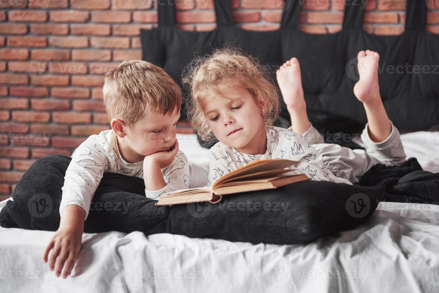 zwei Kinder liegen auf einem großen Bett und lesen ein interessantes Buch. sie tragen den gleichen Pyjama foto