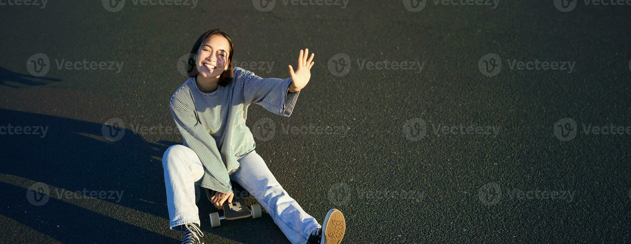 positiv Koreanisch Mädchen Abdeckungen ihr Gesicht von Sonnenlicht, sitzt auf Skateboard und lächelt glücklich foto