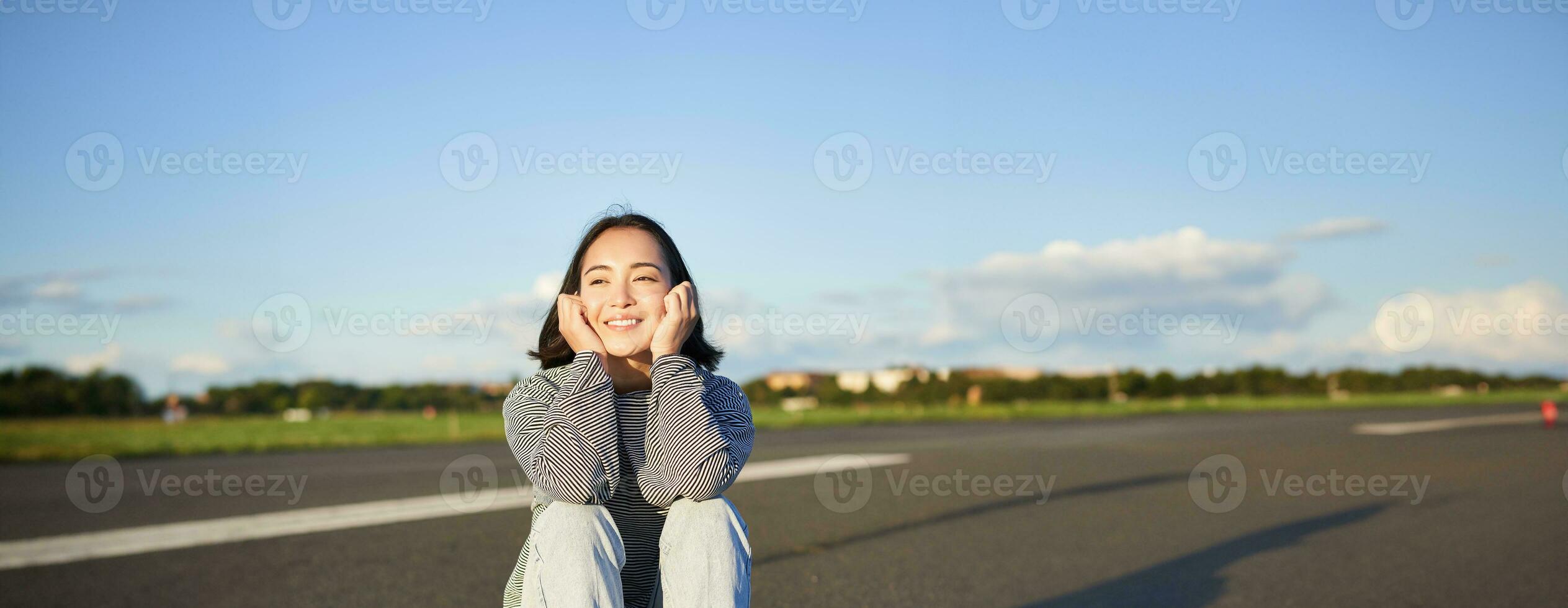 Vertikale Schuss von asiatisch Mädchen Skater, sitzt auf ihr Skateboard und lächelt, genießt sonnig Tag, Kreuzfahrt auf Longboard auf leeren Straße draußen foto