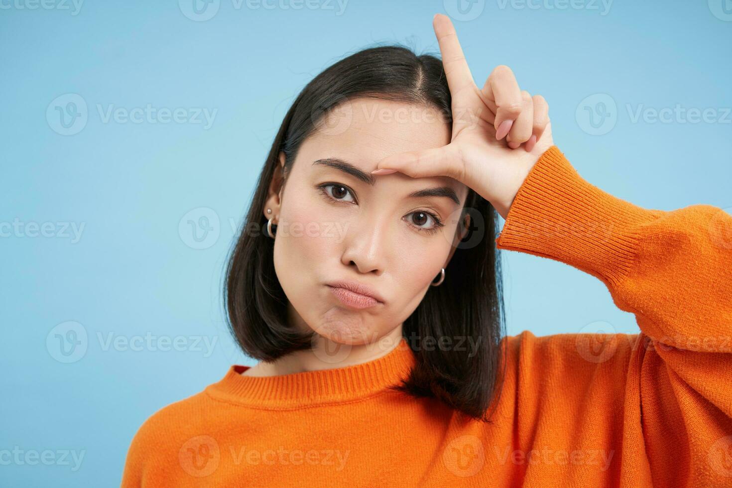 schließen oben von frech Koreanisch Mädchen, zeigt an Brief Ich, Verlierer Symbol auf Stirn, steht Über Blau Studio Hintergrund foto