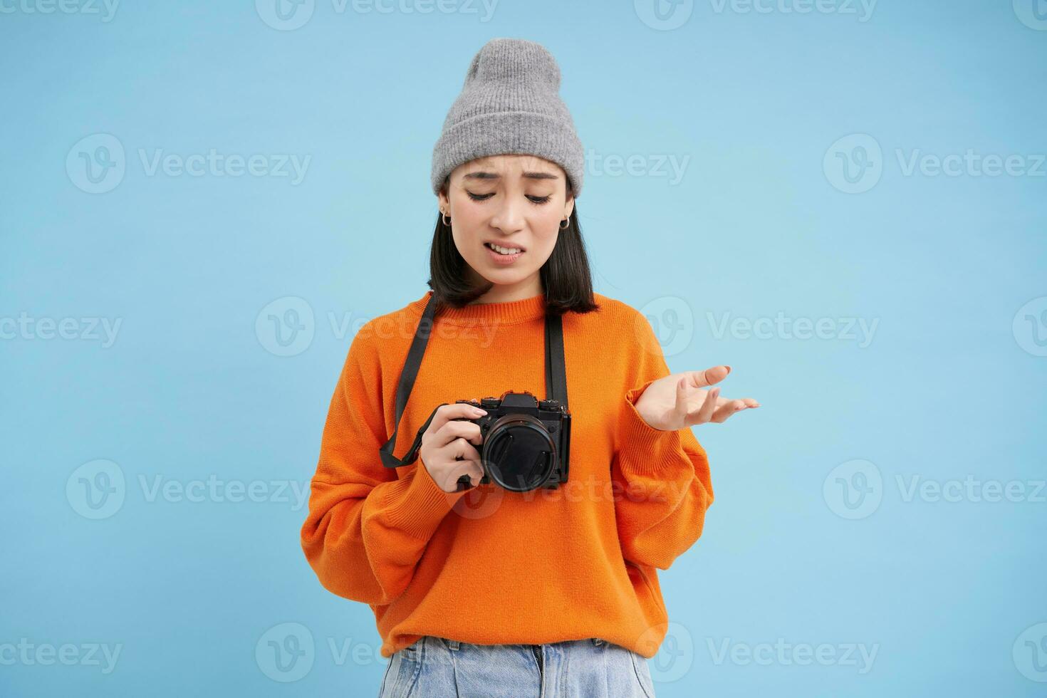 Porträt von asiatisch Frau im Hut, halten Digital Kamera mit verwirrt Gesicht, unprofessionell Fotograf nicht kennt Wie zu nehmen Bilder auf Digicam, Blau Hintergrund foto