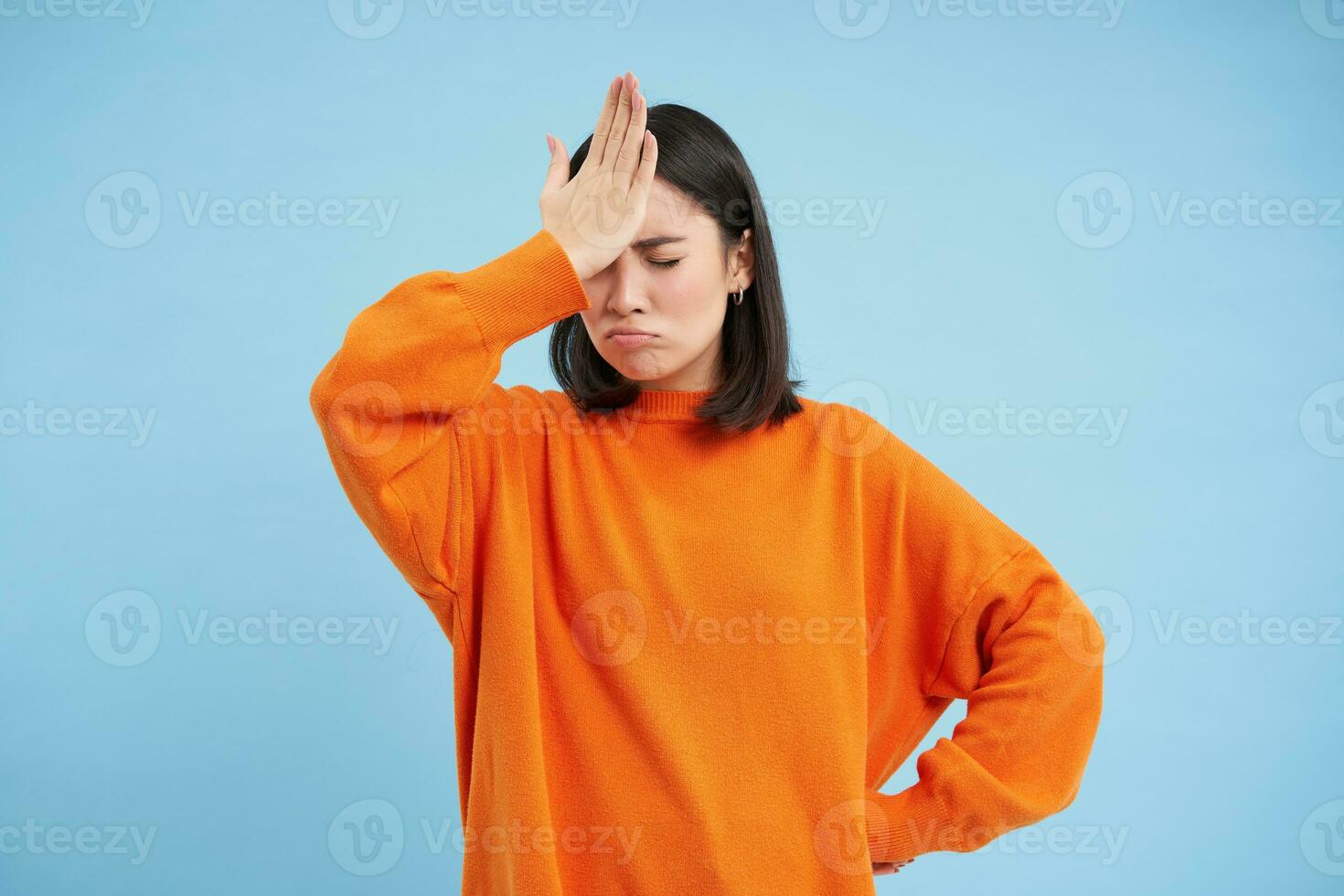 Epos scheitern. enttäuscht asiatisch Frau Ohrfeigen ihr Stirn mit verärgert Gesicht, vergessen etw, verärgert, steht Über Blau Hintergrund im Orange Sweatshirt foto