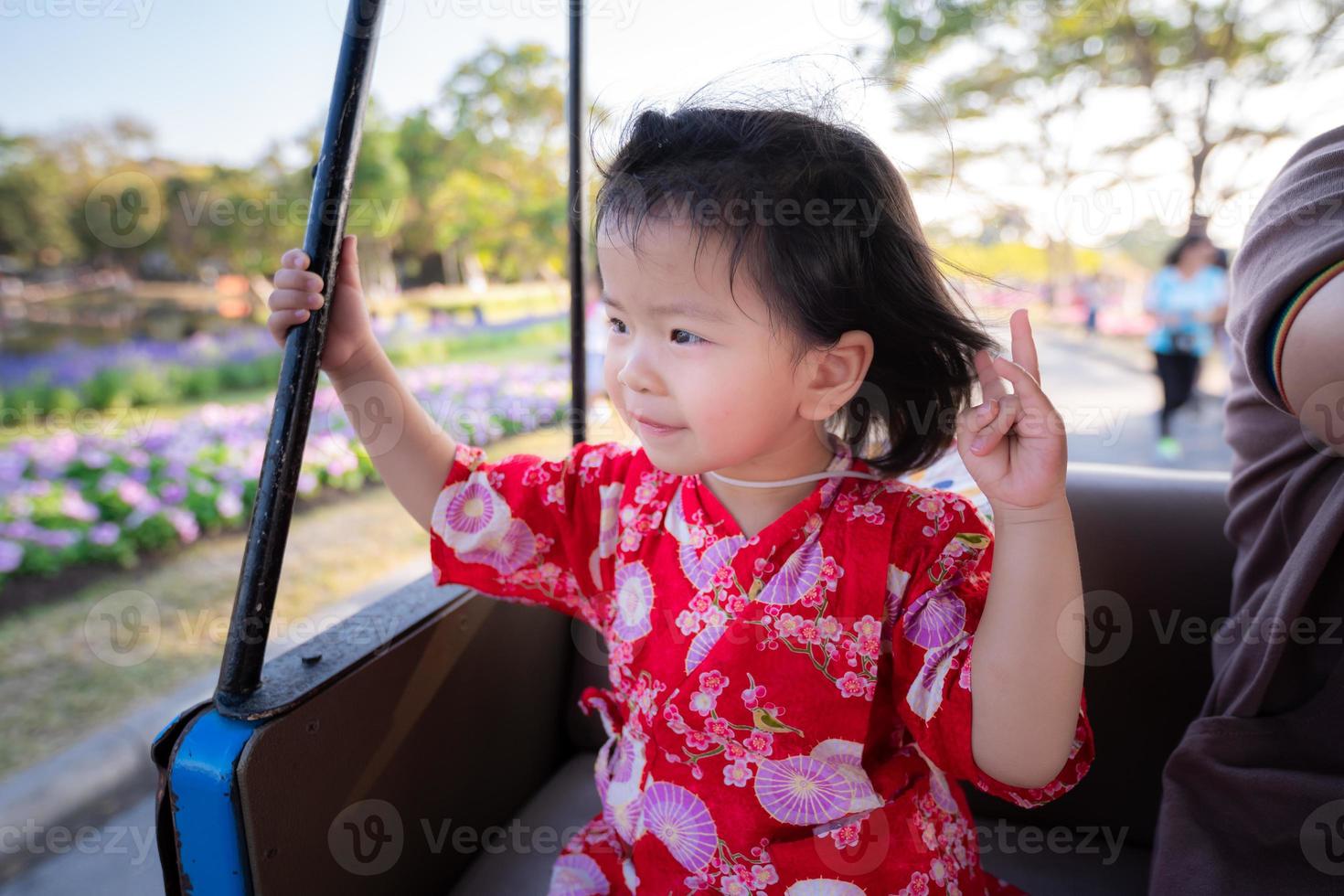 Kopfschuss glückliches Mädchen saß mit süßem Lächeln auf einer Rikscha im Park. Kind in einem traditionellen roten japanischen Kleid sieht an einem schönen Wintertag einen Blumengarten an. süßes smileykind alter 2 jahre alt. foto