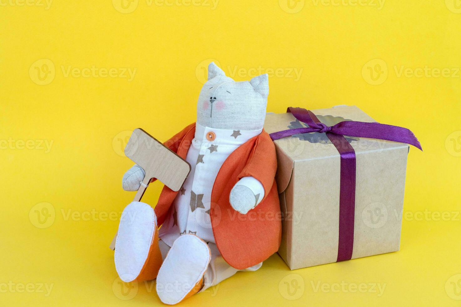 ein Sanft Spielzeug Katze mit ein hölzern Zeichen mit ein Platz zum Text und ein Geschenk Box auf ein Gelb Hintergrund. foto