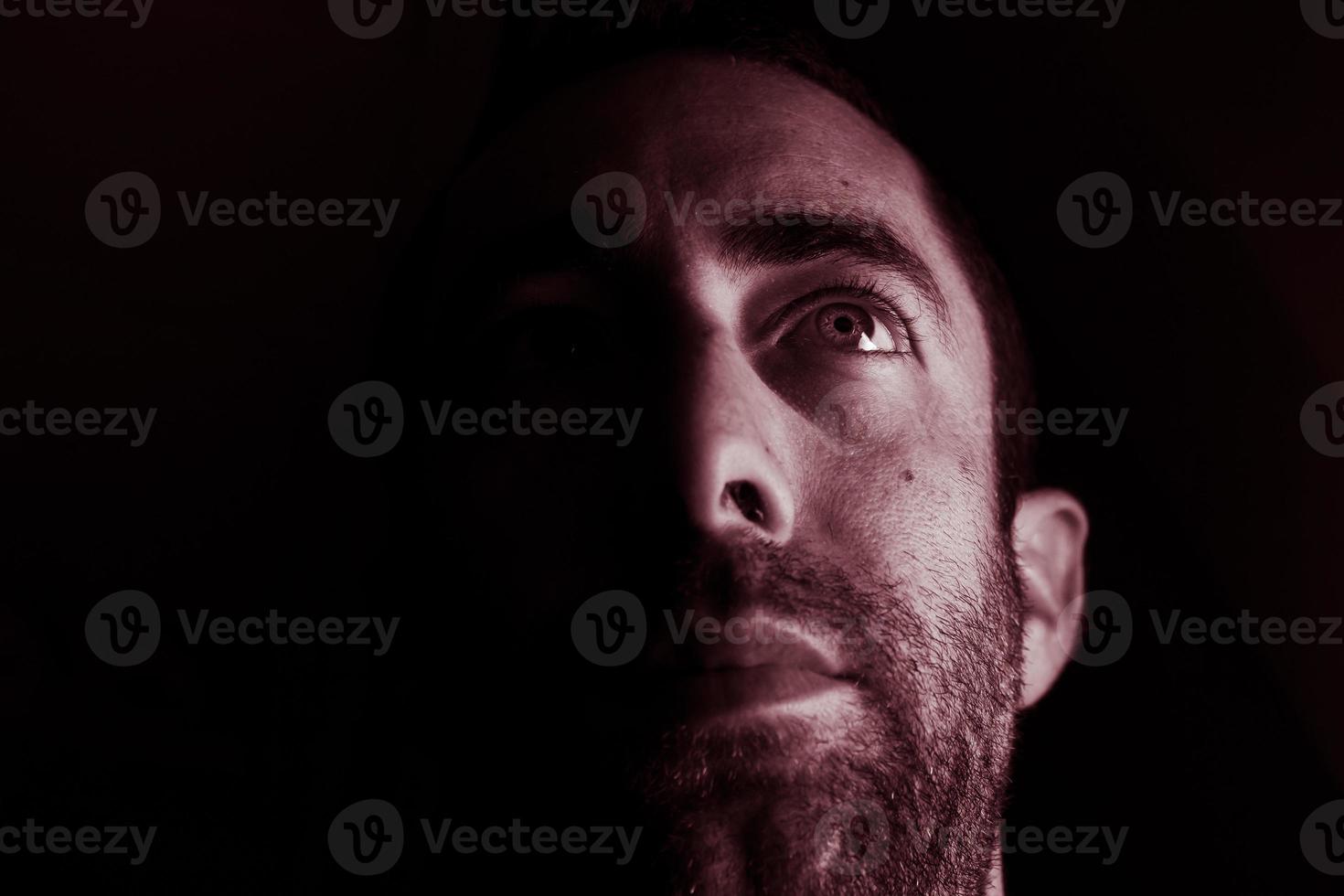 Dramatisches Porträt eines jungen Mannes, halb ausdrucksloses Gesicht im tiefen Schatten, der isoliert auf schwarzem Hintergrund nach oben schaut foto