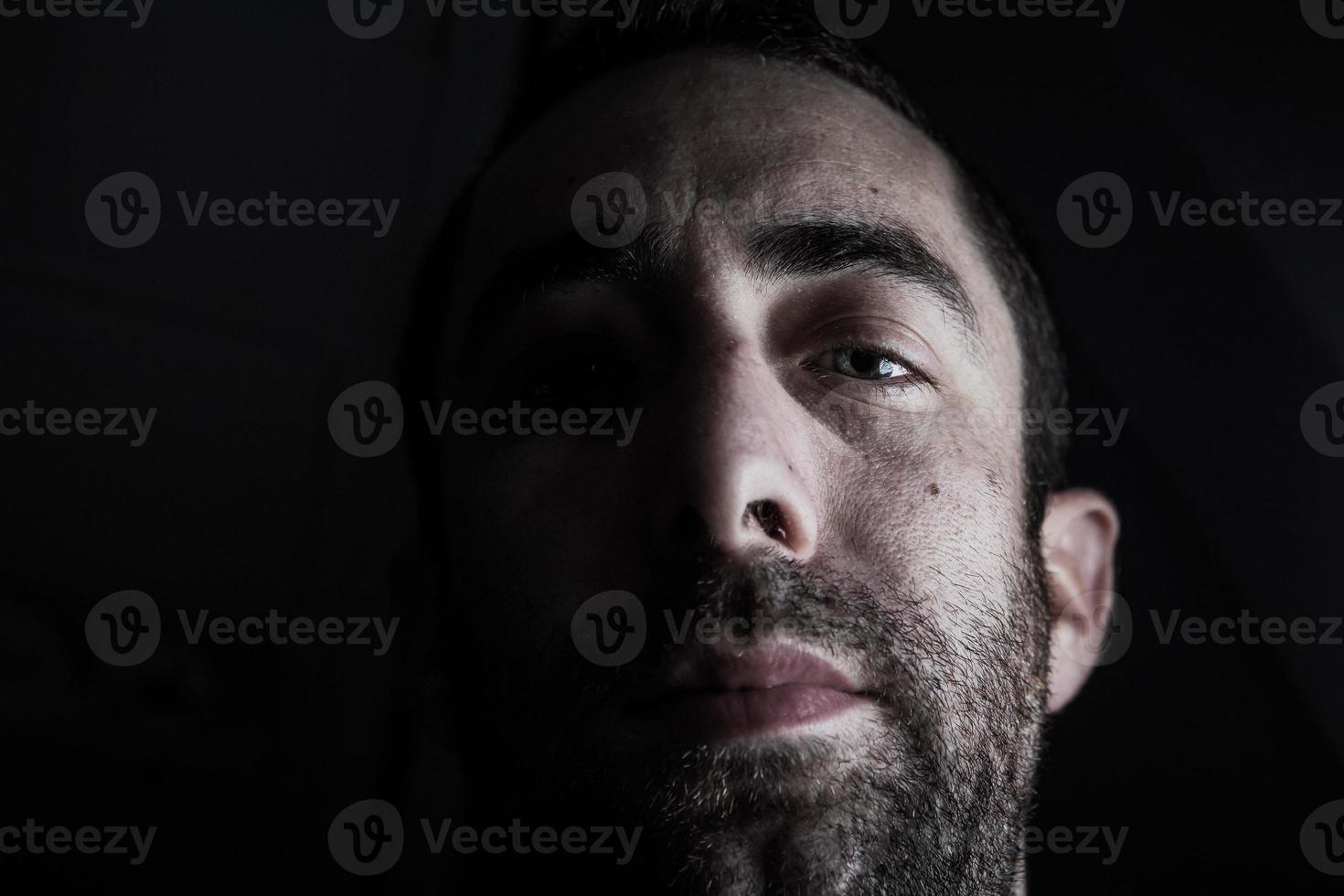 Dramatisches Porträt eines jungen Mannes, halb ausdrucksloses Gesicht im tiefen Schatten, der die Kamera einzeln auf schwarzem Hintergrund betrachtet foto