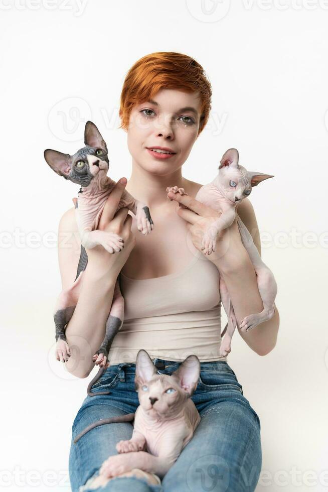 Rothaarige jung Frau halten zwei Sphynx Katze im Hände und einer Kätzchen Lügen auf ihr Beine. ziemlich Frau mit kurz Haar im T-Shirt und Jeans suchen Kamera, Lügen Nieder auf Weiß Hintergrund. Teil von Serie foto