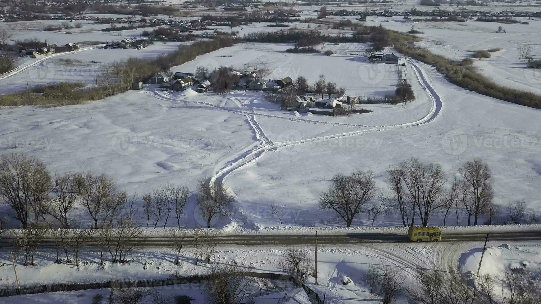 Antenne von das Winter Straße mit ein Bus Reiten entlang das klein Dorf bedeckt durch Schnee. Schuss. Winter Landschaft mit hölzern Häuser, Straße und ziehen um Gelb Bus. foto