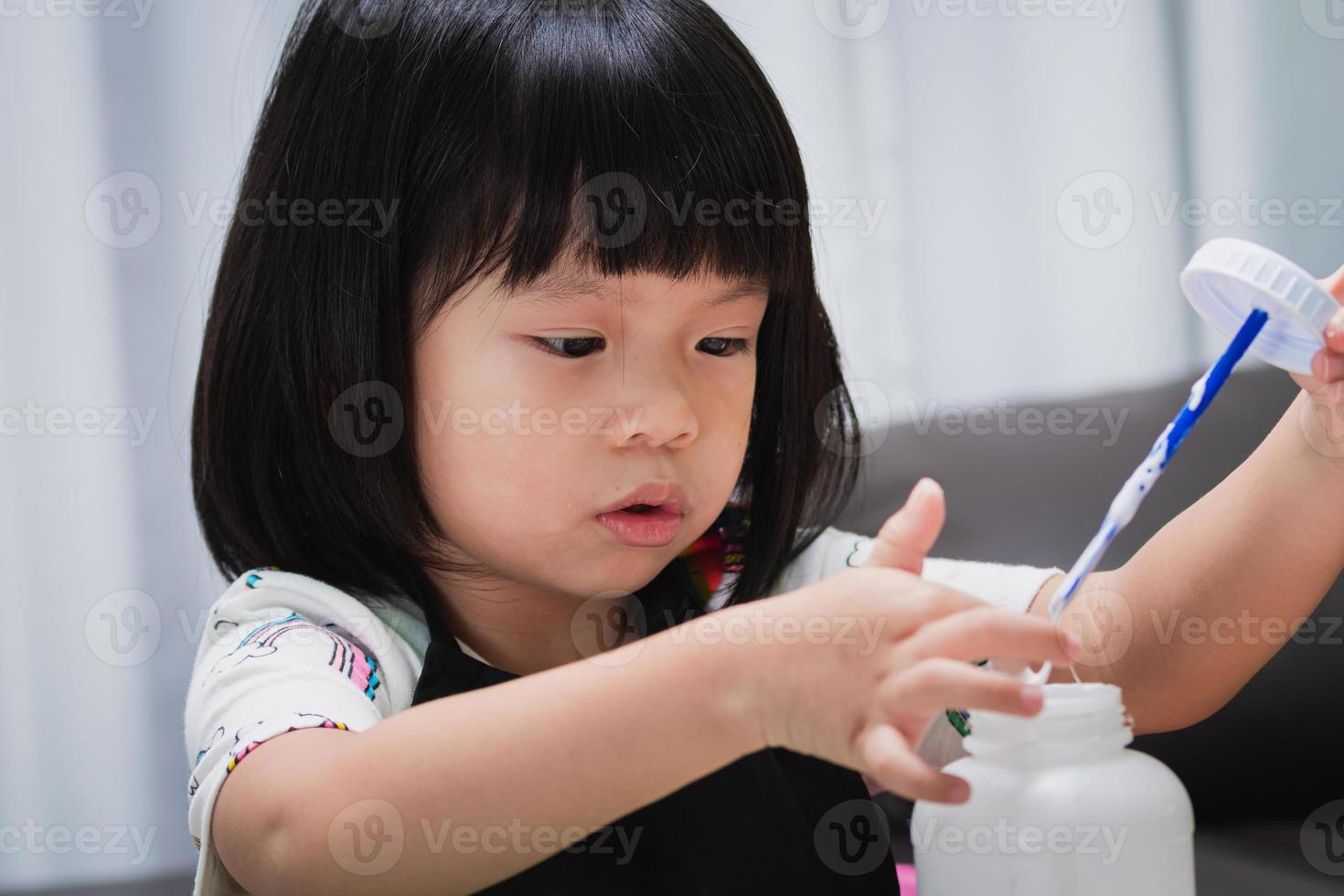 Closeup Gesicht Mädchen lernen Handwerk im Klassenzimmer. Kindergartenschüler, der Kleber berührt, um Erfindung zu machen. Vorschulkind mit lustigem DIY-Projekt. Baby im Alter von 4-5 Jahren mit schwarzer Schürze. foto