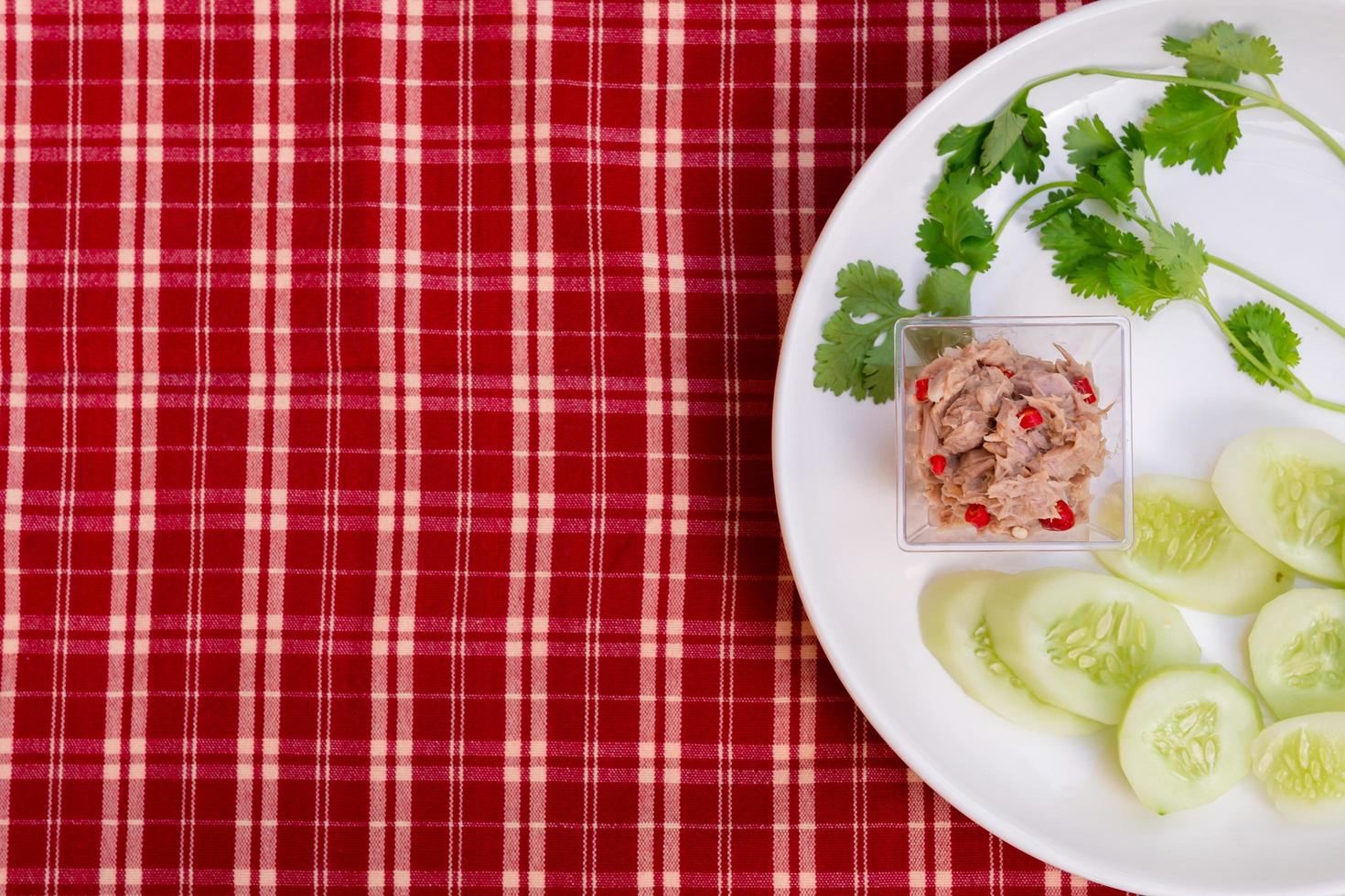 würziger Salat mit Mini-Thunfisch und Koriander und Gurkenscheiben auf einem weißen Teller. roter Stoffhintergrund, Gingham. foto