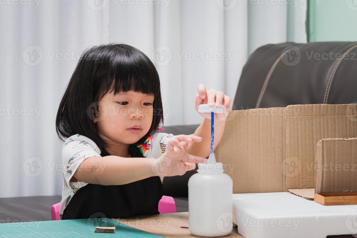4-5 Jahre altes Mädchen baut ein Haus aus Kartons und Styropor. Kind verwendet Klebstoff, um kleine Teile vorsichtig aufzutragen, um sie in ihrem Haus zu montieren. Kind hat wirklich Spaß daran zu lernen und es zu tun. foto