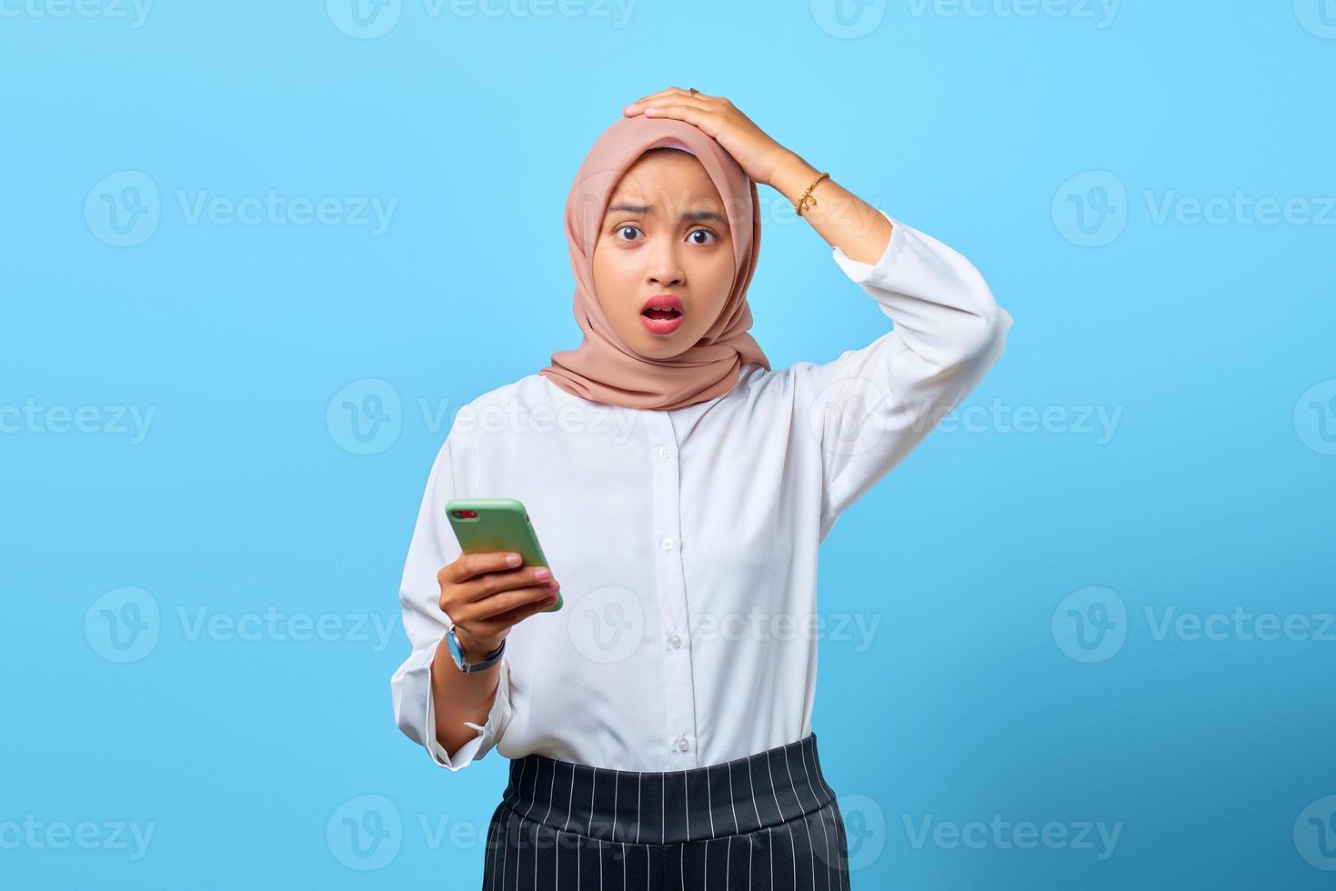 Porträt einer überraschten jungen asiatischen Frau, die Handy mit der Hand auf dem Kopf über blauem Hintergrund benutzt foto