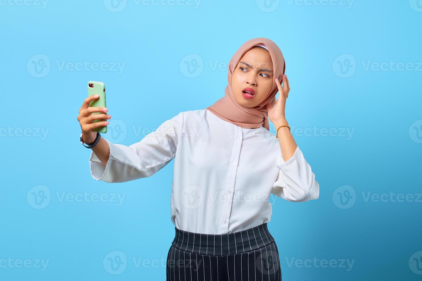 Porträt einer schockierten jungen Asiatin mit offenem Mund mit Handy über blauem Hintergrund foto