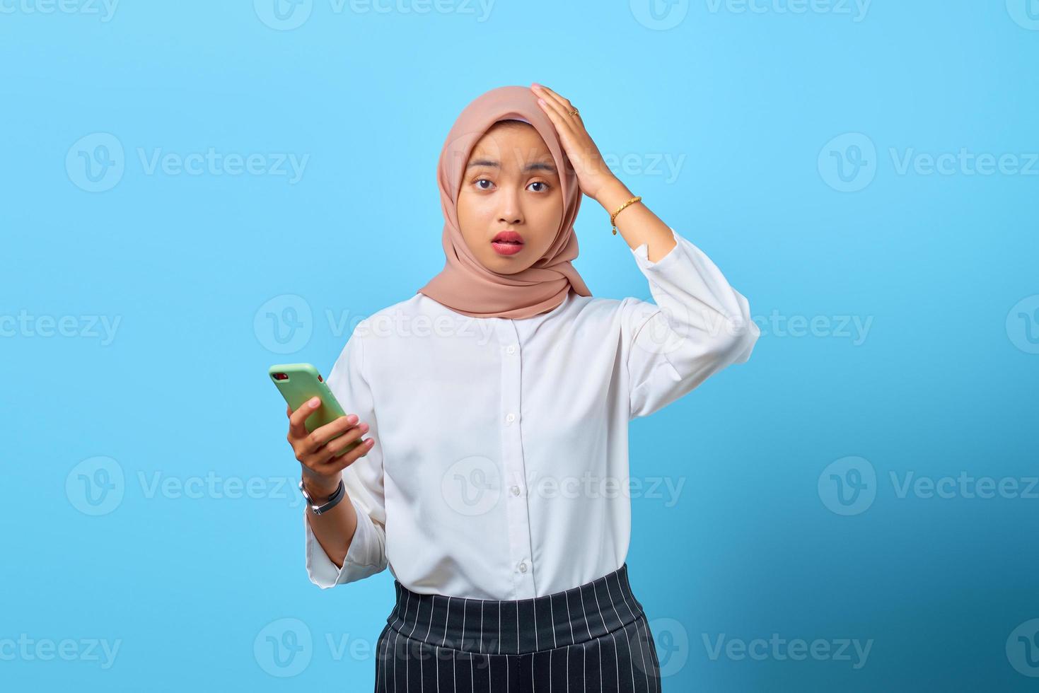 Porträt einer überraschten jungen asiatischen Frau, die Handy mit der Hand auf dem Kopf über blauem Hintergrund benutzt foto