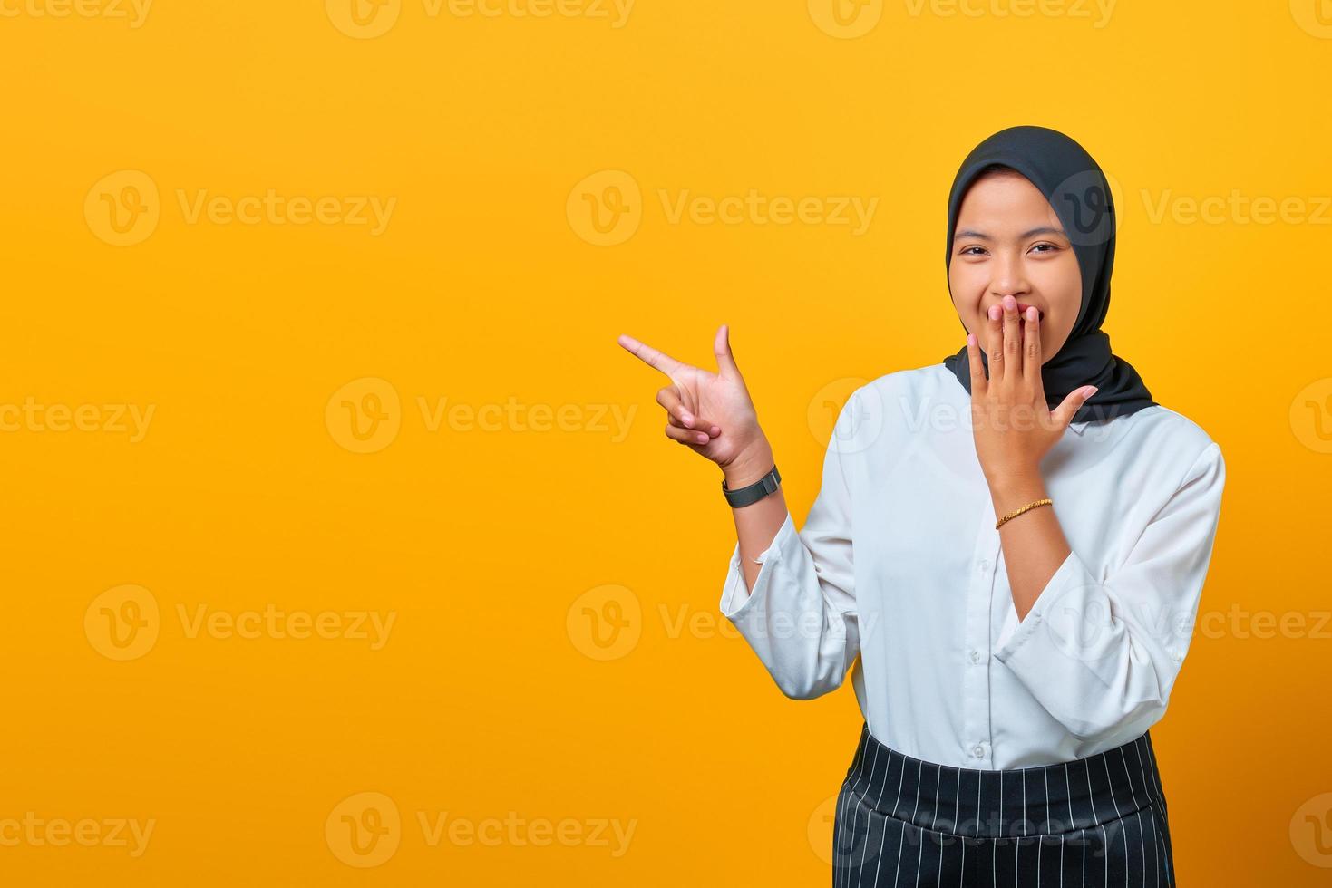 lächelnde junge asiatische frau, die mit den fingern auf den kopierraum zeigt und die kamera auf gelbem hintergrund schaut foto