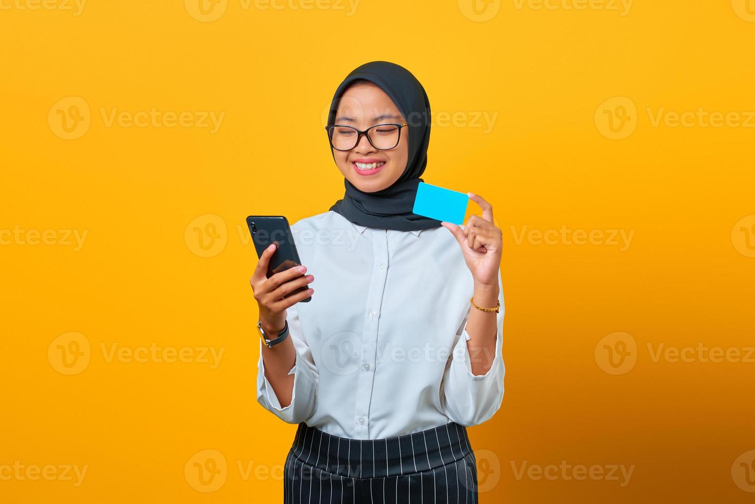 fröhliche junge asiatische frau, die handy und kreditkarte auf gelbem hintergrund hält foto