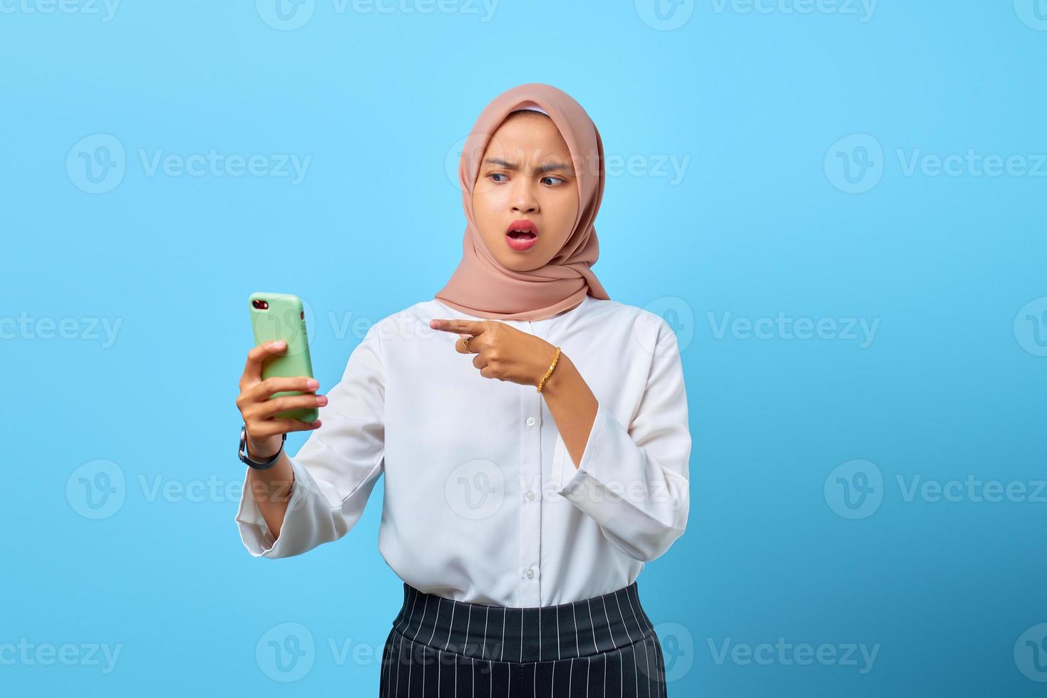 Porträt einer schockierten jungen asiatischen frau, die mit offenem mund auf blauem hintergrund auf handy zeigt foto