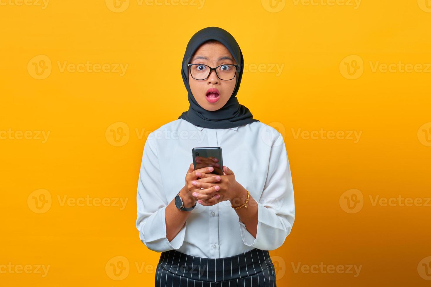 überraschte junge asiatische frau, die handy mit offenem mund auf gelbem hintergrund hält foto