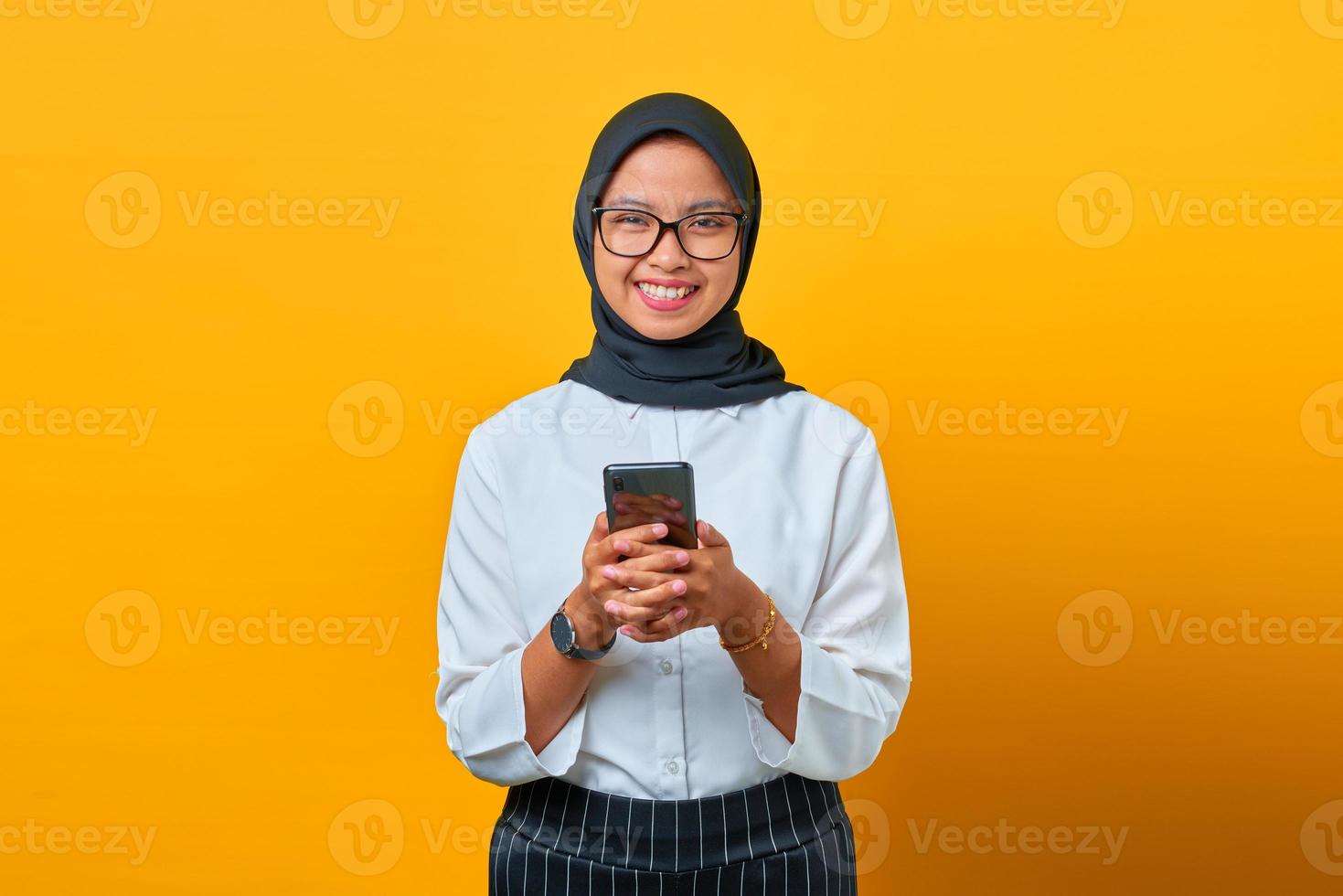 lächelnde junge asiatische frau, die handy hält und kamera auf gelbem hintergrund schaut foto