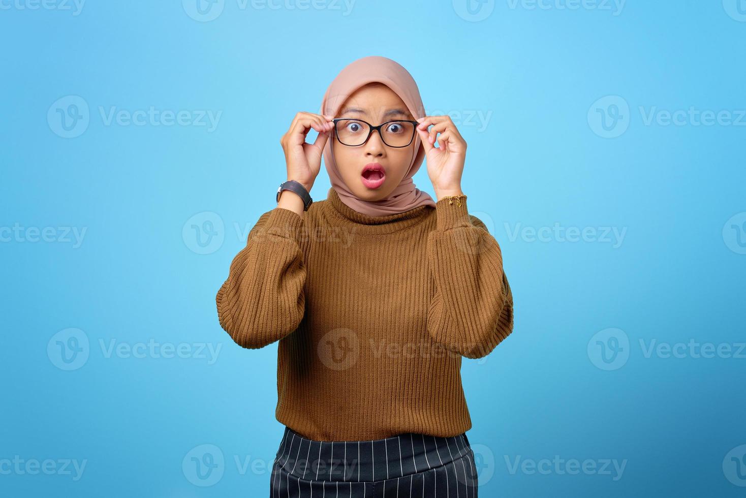 überraschte junge asiatische frau hand auf brille mit offenem mund auf blauem hintergrund foto