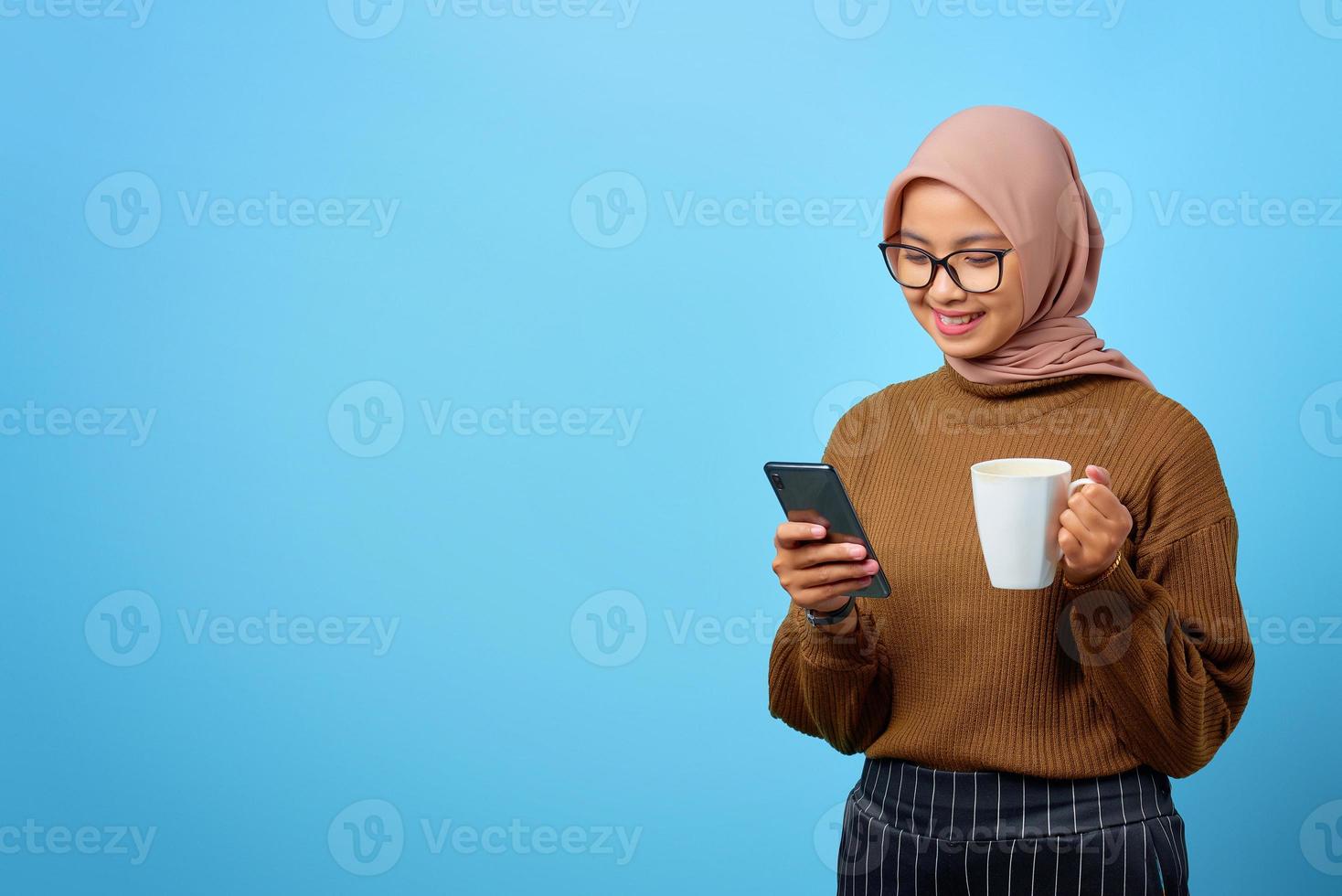 Fröhliche junge asiatische Frau, die sich entspannt, eine Tasse hält und einen Smartphone-Bildschirm auf blauem Hintergrund sieht foto