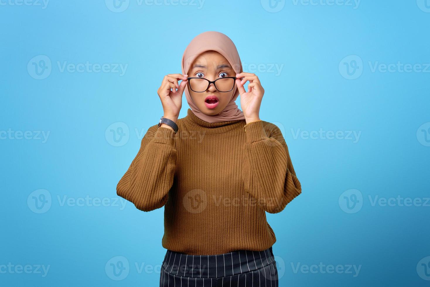 überraschte junge asiatische frau, die brille berührt und kamera auf blauem hintergrund schaut foto