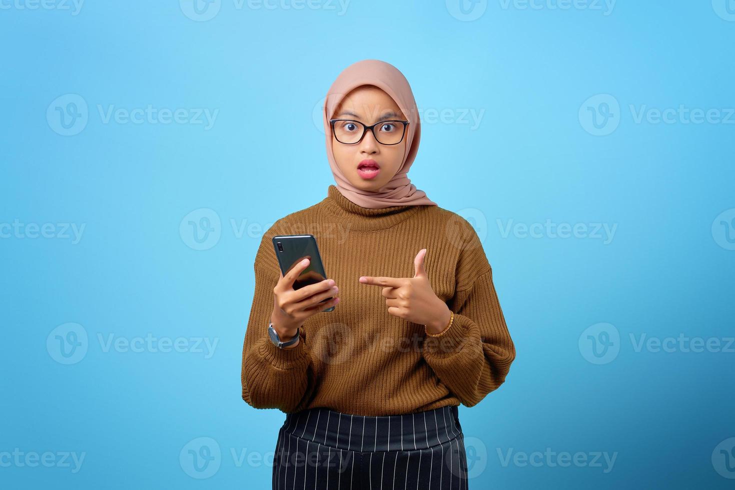 überraschte junge asiatische frau, die mit offenem mund auf blauem hintergrund auf handy zeigt foto