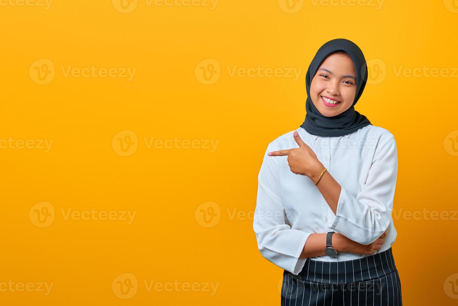 lächelnde junge asiatische frau, die mit den fingern auf den kopierraum zeigt und die kamera auf gelbem hintergrund schaut foto