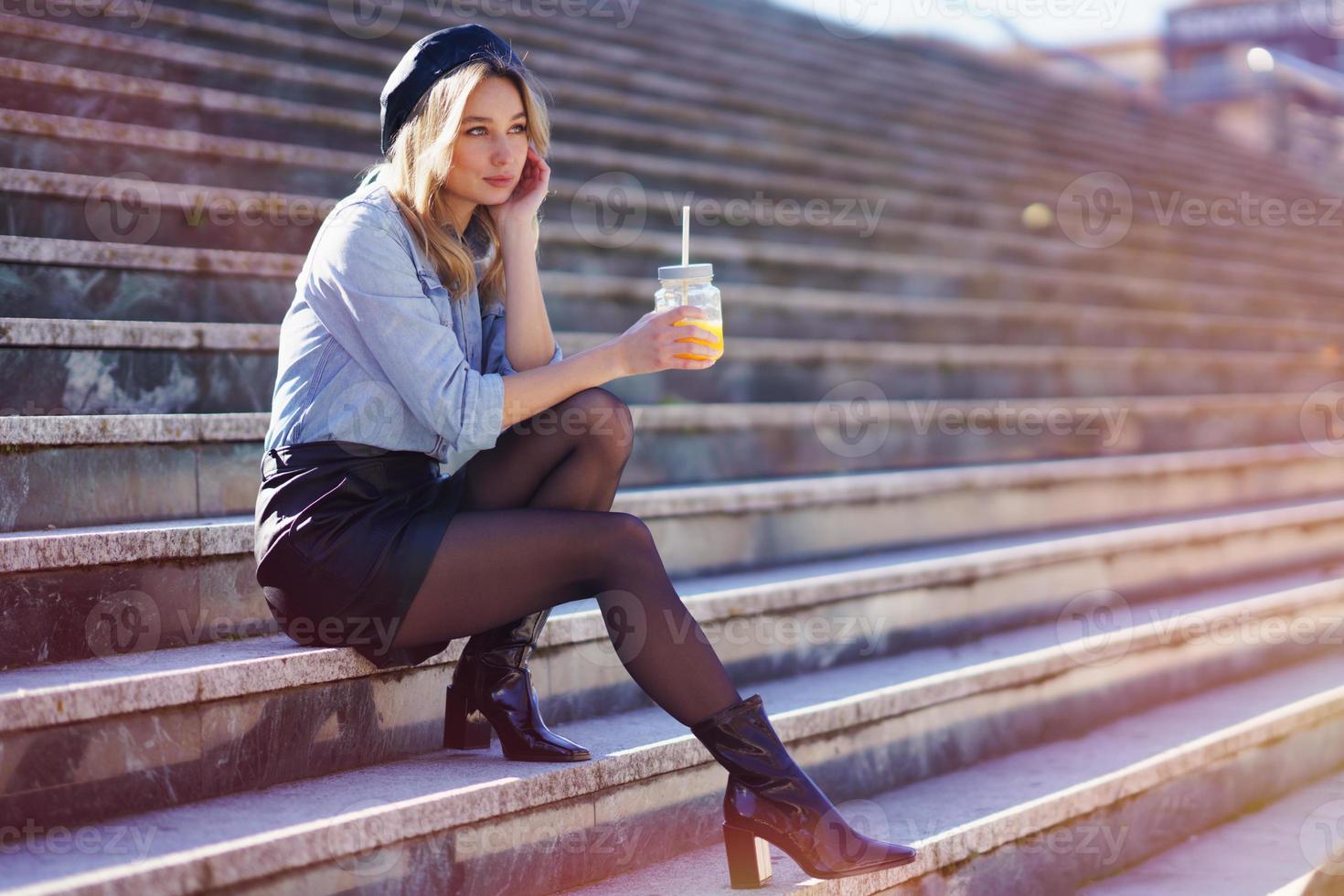 blonde Frau mit Baskenmütze, einen natürlichen Orangensaft in einem Kristallglas trinkend, auf einigen Stufen sitzend. foto