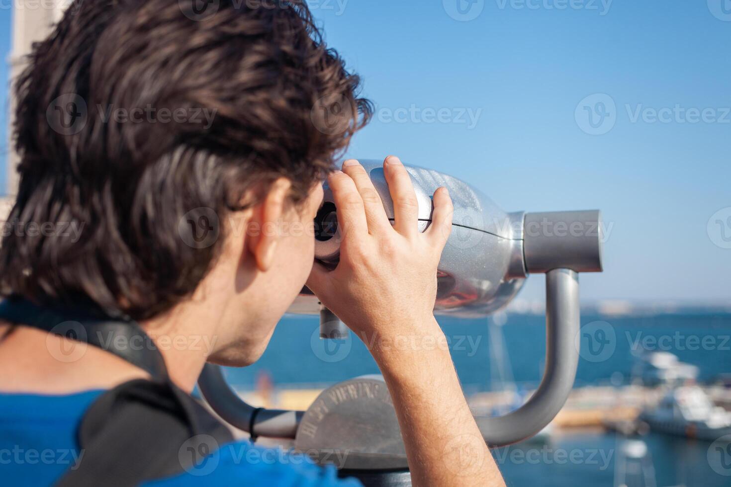 Mann sieht aus durch stationär Fernglas. Binoskop auf Überwachung Deck in der Nähe von Meer. Seehafen im Odessa. teuer Yachten auf das Seebrücke. kommerziell Maschine, Verkauf Geschäft. foto