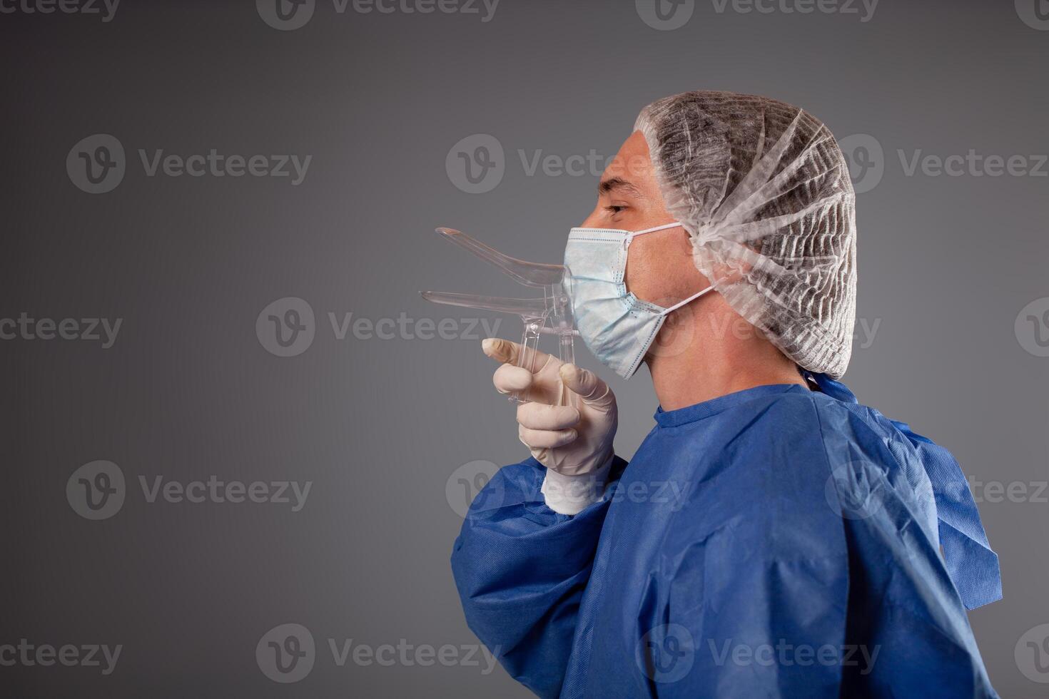 ein Gynäkologe mit ein Venus Spiegel im seine Hände. gynäkologisch Zubehör, Bürste, Stock. ein Arzt im ein medizinisch Maske, ein Kleid, Handschuhe, mit ein gynäkologisch Spiegel auf ein grau Hintergrund. foto