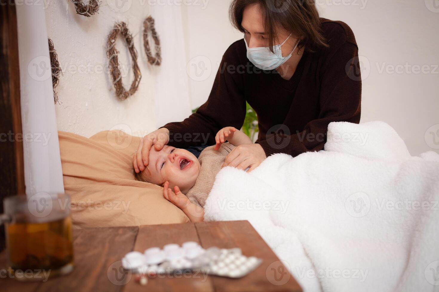 das Kind beim Zuhause ist krank, schreiend und leiden. Papa im ein Maske prüft seine Tochter Temperatur. ein Kind habe krank, Coronavirus, Covid, kalt, Grippe. ein Mädchen auf das Hintergrund von Pillen, Quarantäne foto