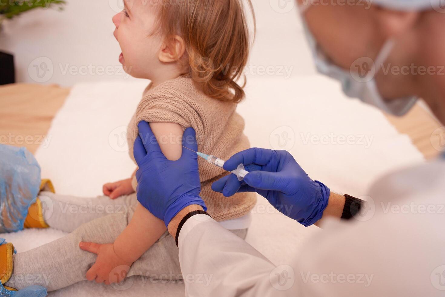 das Arzt impft das Kind gegen Coronavirus. das Kind ist Weinen und besorgt. ein Mann im ein Kleid, Hut, Maske und Handschuhe macht ein Injektion zu das Kind. Zuhause Quarantäne foto