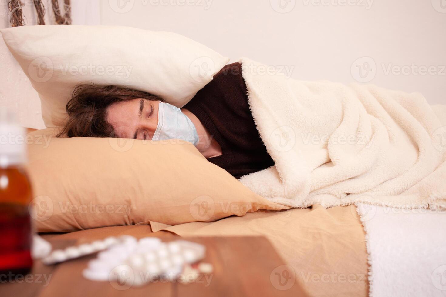 ein krank Mann im ein medizinisch Maske gegen das Hintergrund von Tablets. Zuhause Quarantäne, Coronavirus, Covid. das Mann ist bedeckt mit ein Kissen und schlafend foto