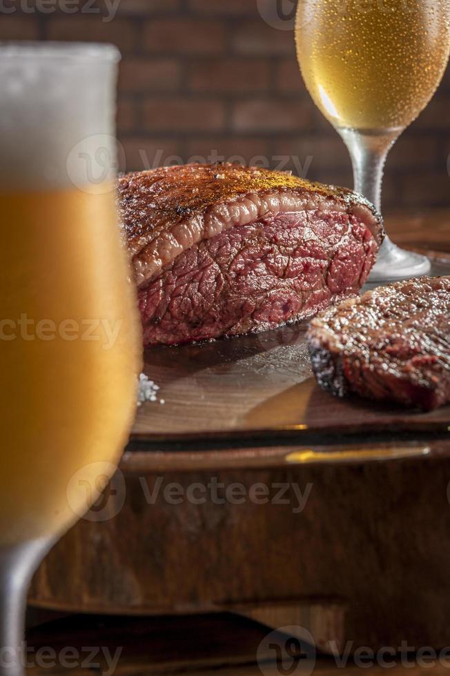 Gegrilltes Rumpsteak in Scheiben geschnitten mit zwei Gläsern Bier auf Holzbrett. Marmorfleisch Rindfleisch - brasilianischer Picanha. foto