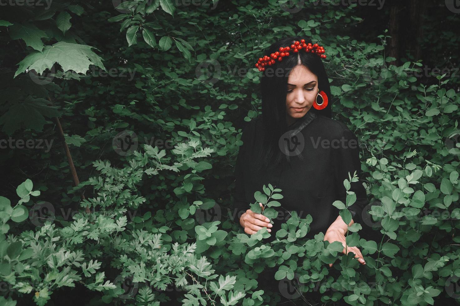 Schönes Mädchen in schwarzem Kleid und roten Dekorationen, das vor dem Hintergrund des Waldes einen Zweig in der Hand hält. Platz für Ihre SMS oder Werbeinhalte foto