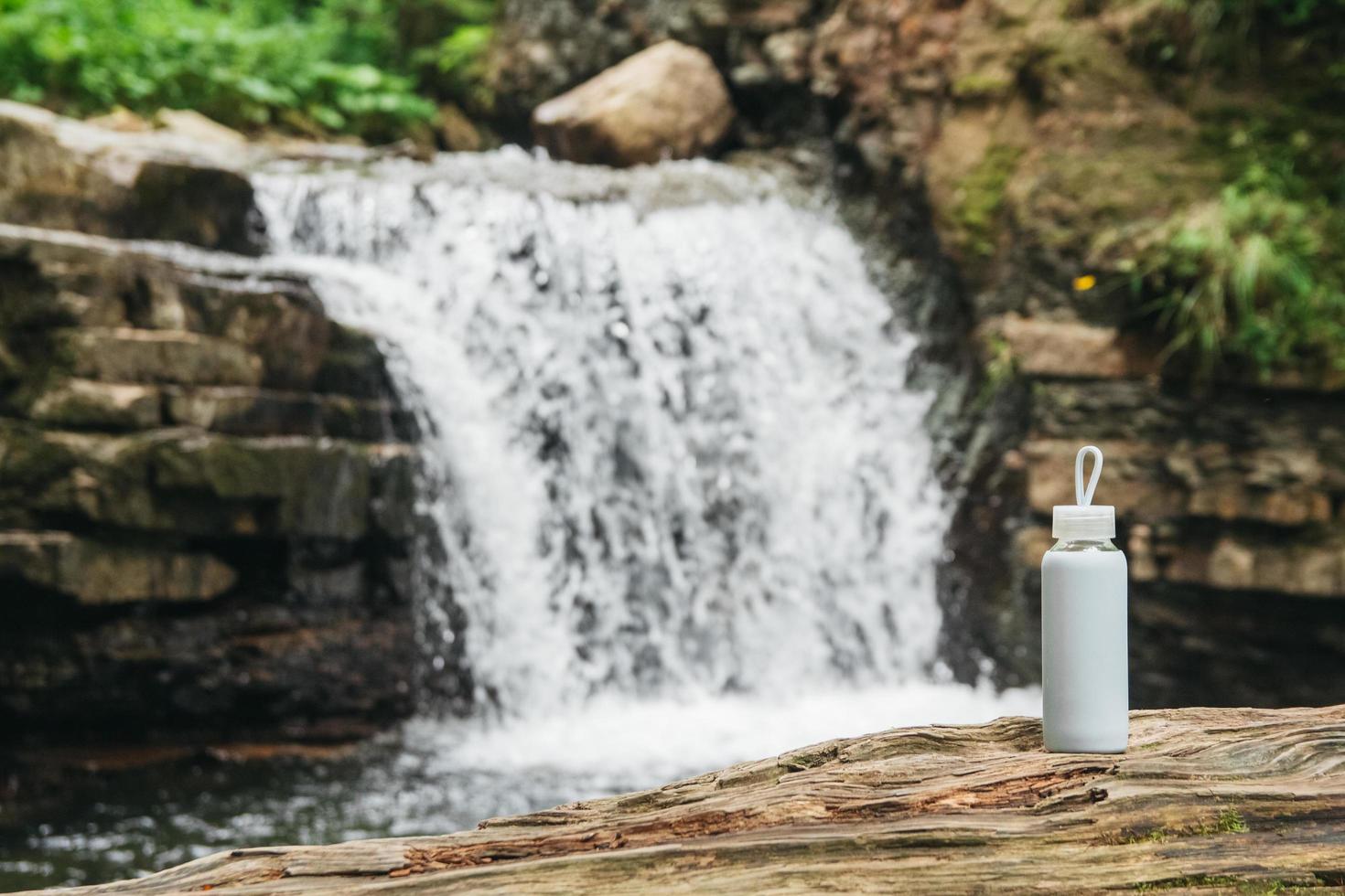 weiße Flasche mit Wasser, die auf einem Holzstamm vor dem Hintergrund eines Flusses und eines Wasserfalls steht. das Konzept des reinen natürlichen Trinkwassers foto