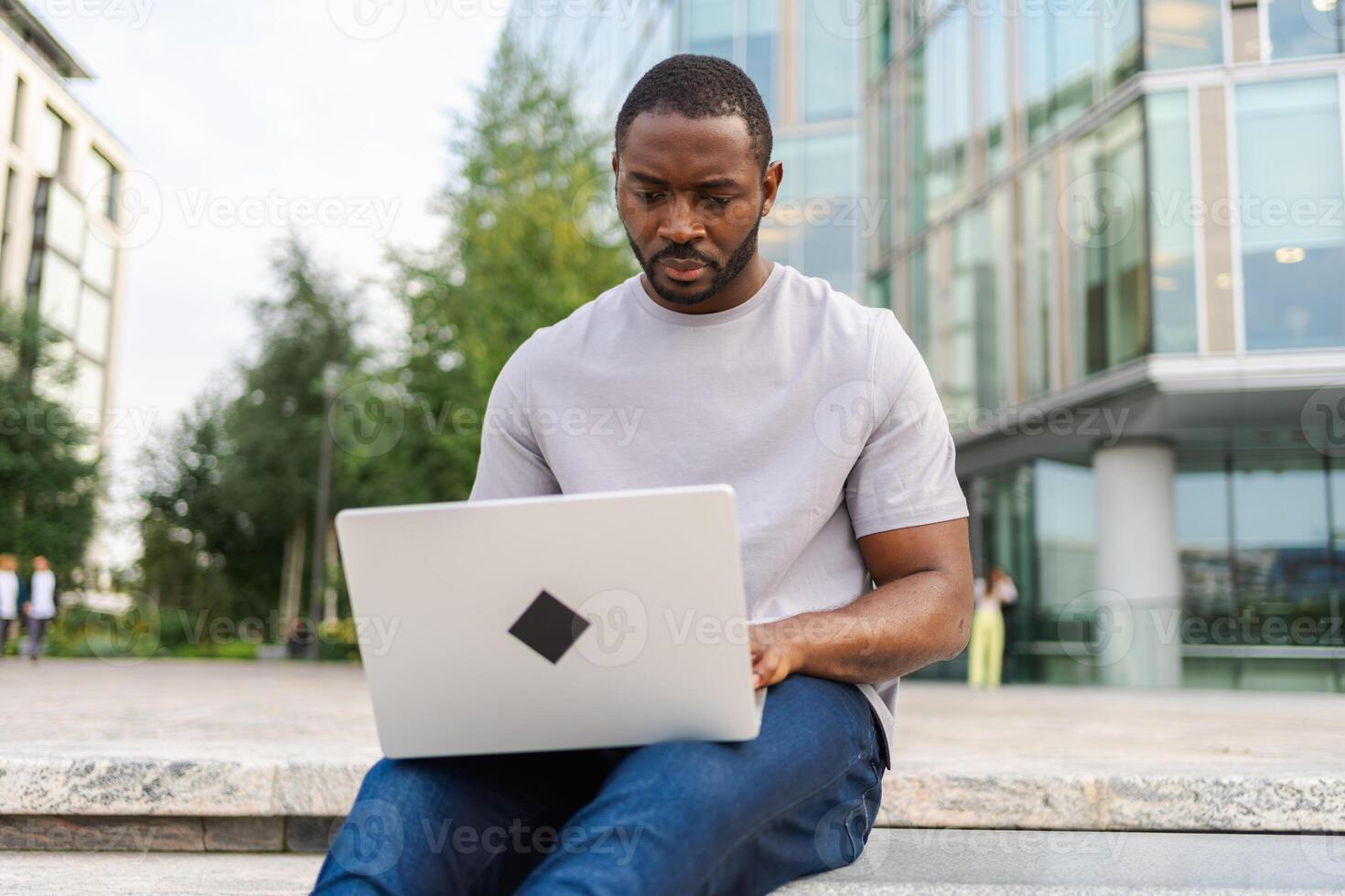 afrikanisch amerikanisch Mann Freiberufler mit Laptop Tippen chatten auf städtisch Straße im Stadt. Kerl haben virtuell Treffen online Plaudern Konferenz. Unternehmer Programmierer Arbeiten auf Laptop draußen. Fernbedienung Arbeit foto