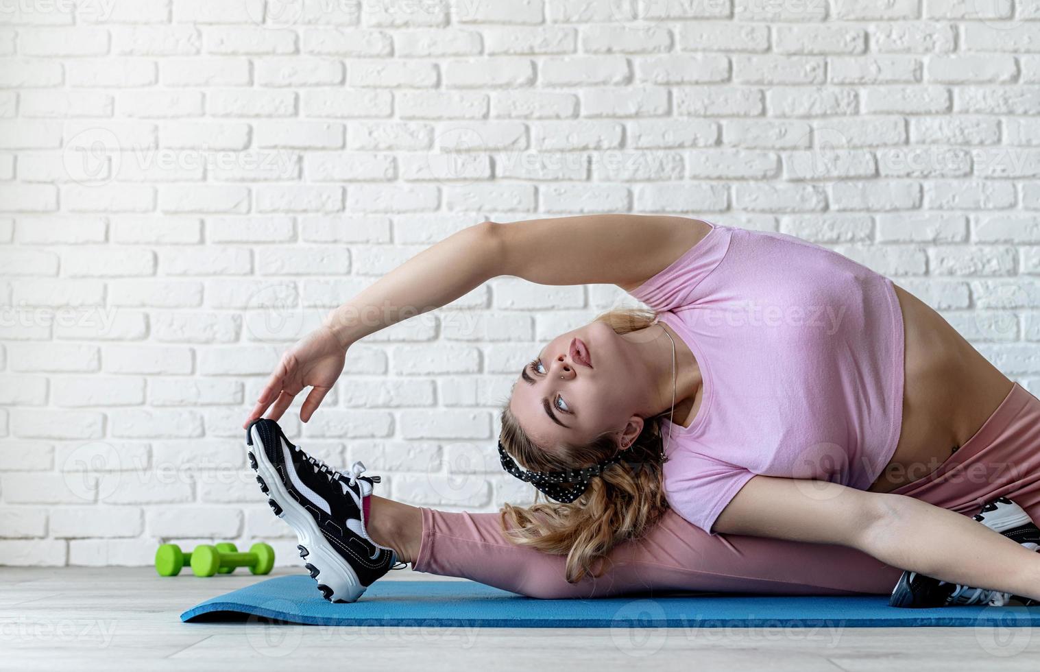 athletische Frau, die sich zu Hause auf einer Fitnessmatte am weißen Backsteinmauerhintergrund ausdehnt foto