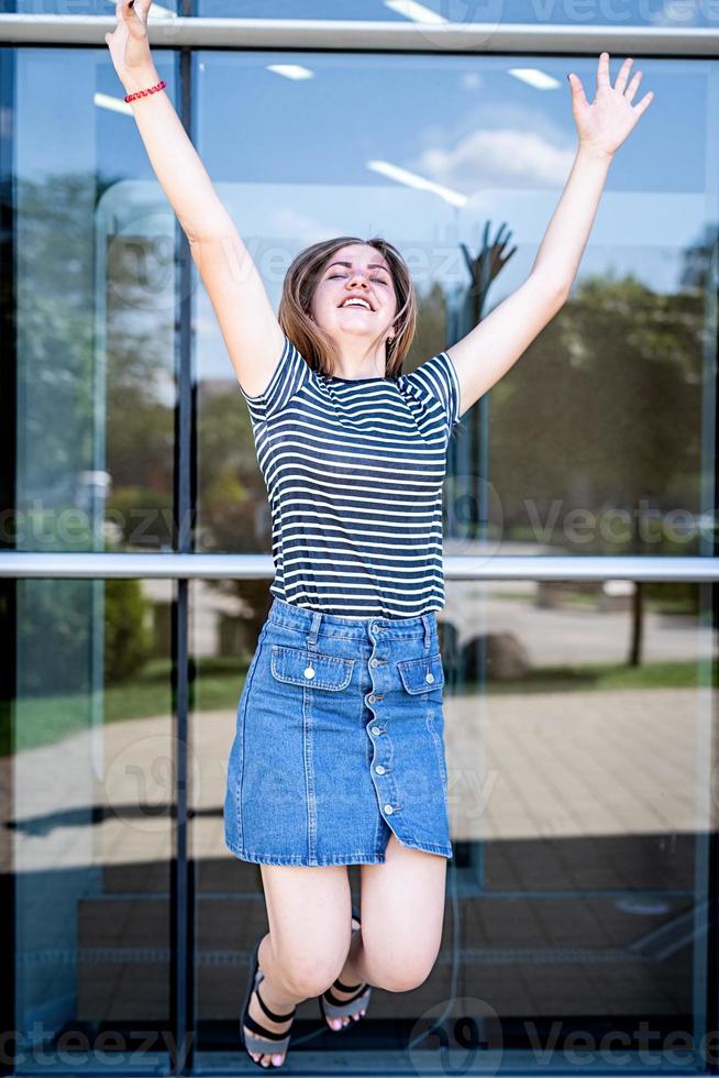 Ganzaufnahme einer fröhlichen jungen kaukasischen Frau, die über den städtischen Hintergrund des Spiegels springt und feiert foto