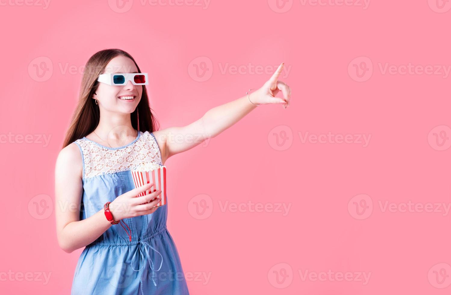 weibliches Teenager-Mädchen mit 3D-Brille isst Popcorn und zeigt mit dem Finger isoliert auf rosa Hintergrund foto