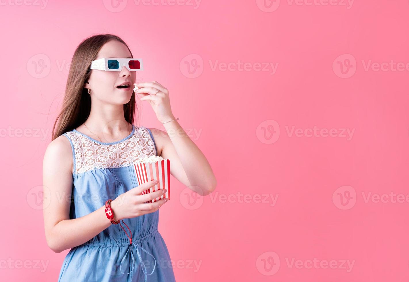 aufgeregtes weibliches Teenagermädchen mit 3D-Brille, das Popcorn isst, isoliert auf rosa Hintergrund foto