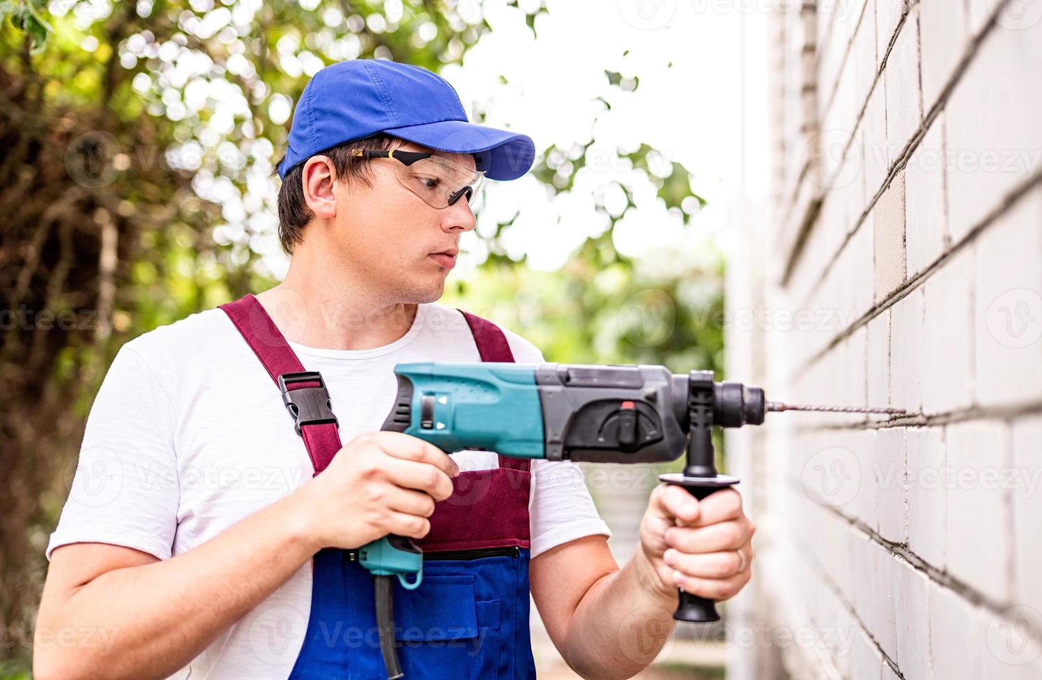 Bauarbeiter in Schutzbrille und Uniform mit Perforator, der die Wand im Freien bohrt. Mann mit Bohrer foto