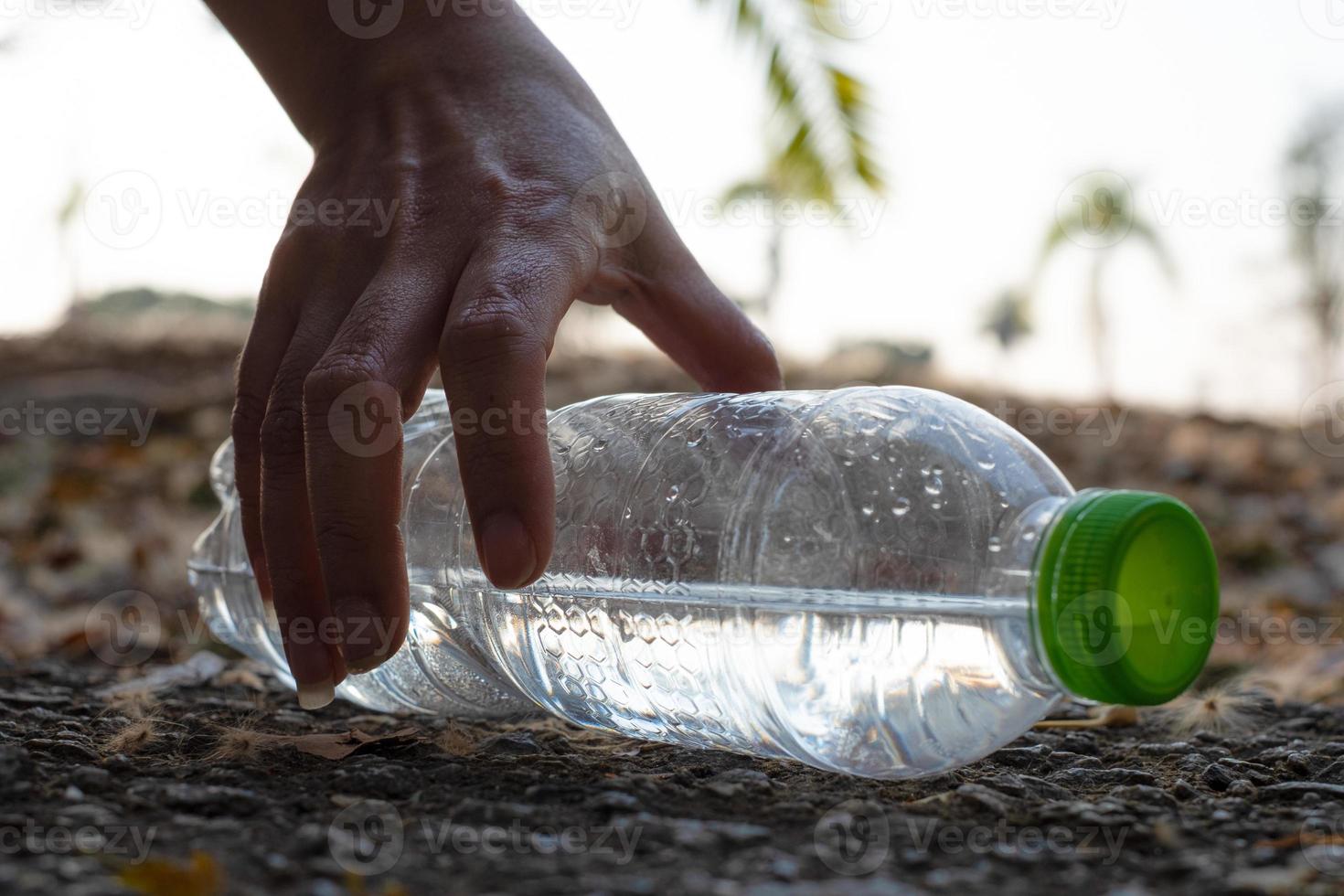 Nahaufnahme von Hand, die klares Plastikflaschenwasser mit einer grünen Kappe auf der Straße im Park auf unscharfem Hintergrund aufhebt, Müll, der außerhalb des Mülleimers zurückgelassen wird foto