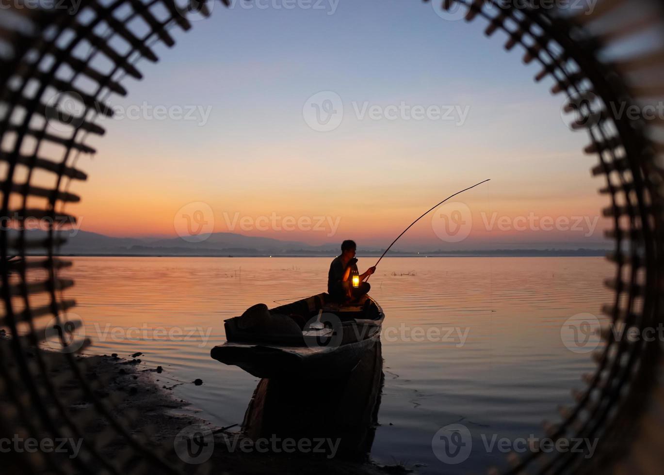 Am Seeufer sitzt ein asiatischer Fischer auf dem Boot und verwendet eine Angelrute, um bei Sonnenaufgang Fische zu fangen foto