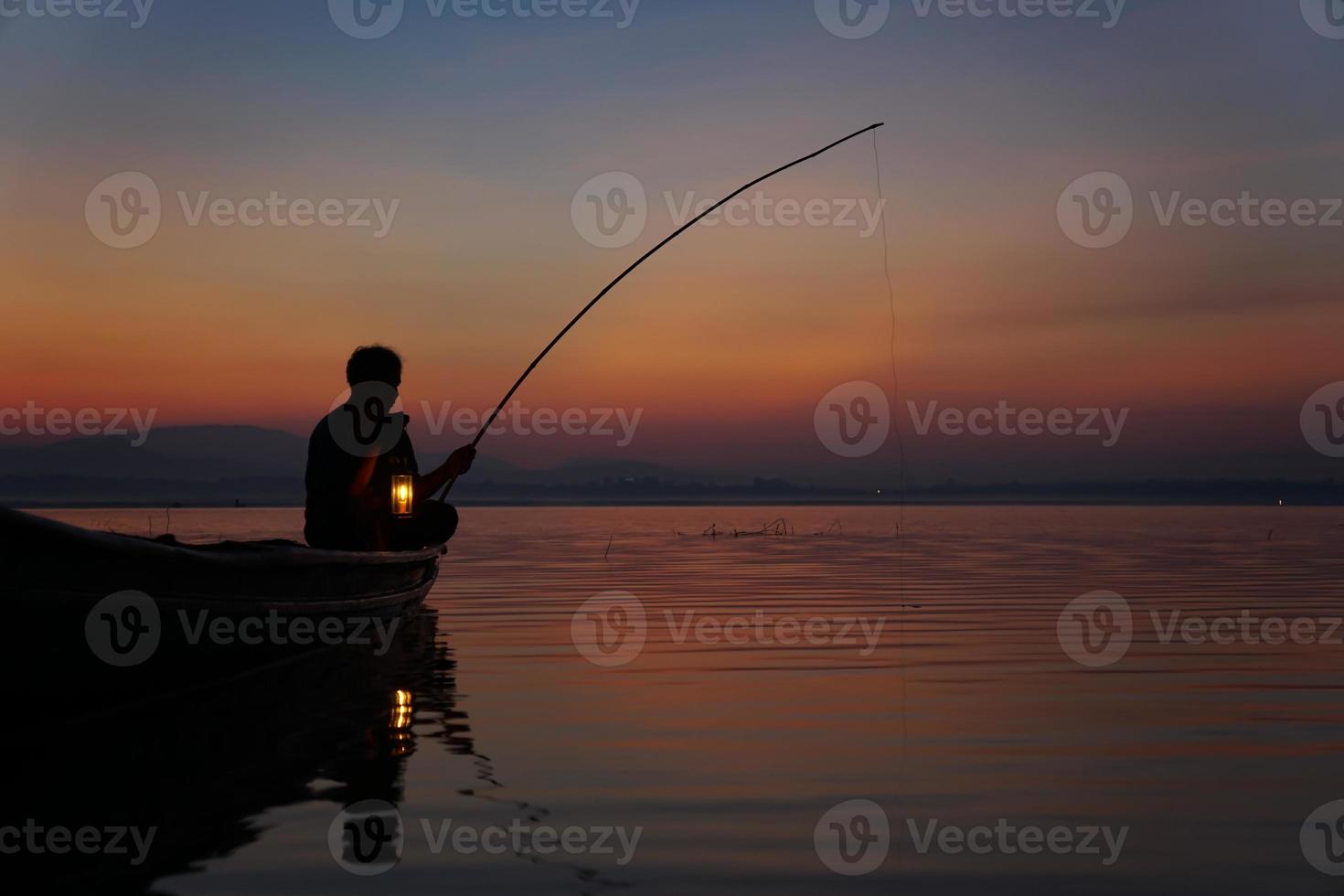 Am Seeufer sitzt ein asiatischer Fischer auf dem Boot und verwendet eine Angelrute, um bei Sonnenaufgang Fische zu fangen foto