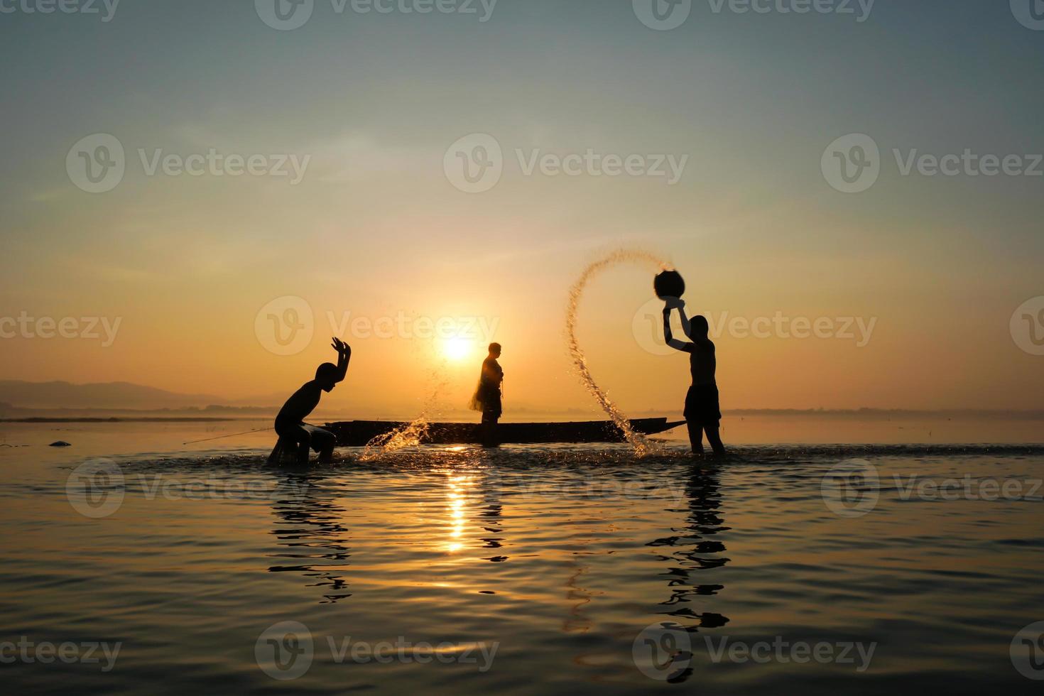Asiatischer Fischer, der auf dem Boot steht und mit Angelrute Fische fängt, während sein Sohn morgens am Seeufer Wasser spritzt? foto