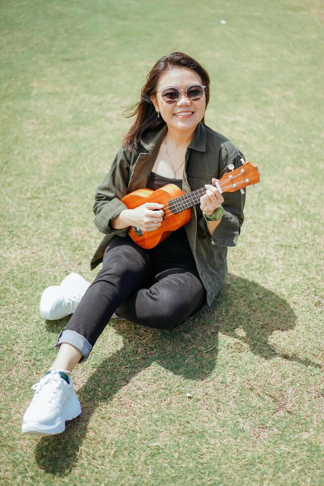 spielen Ukulele von jung schön asiatisch Frau tragen Jacke und schwarz Jeans posieren draußen foto